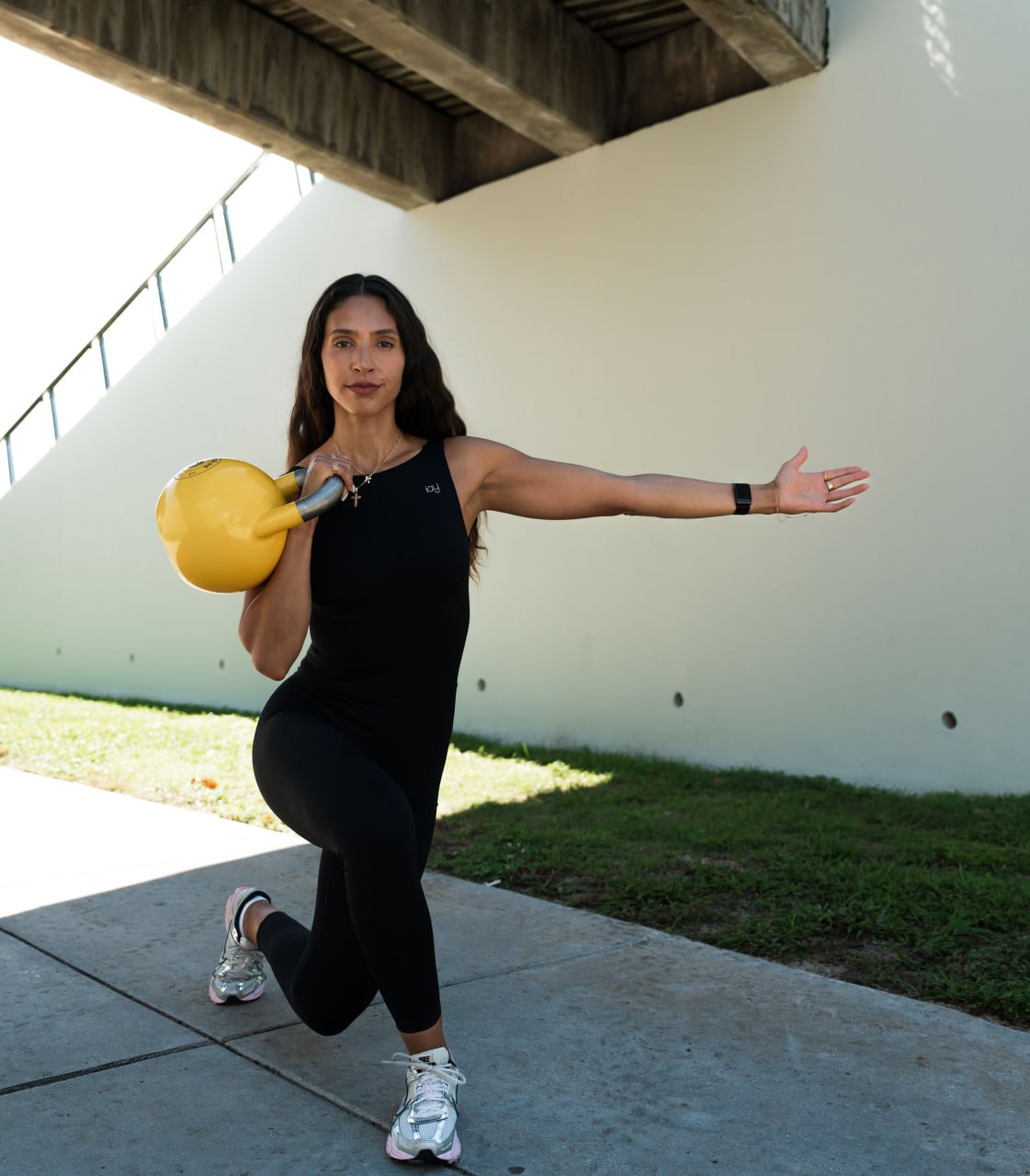 Krizia Valentino holds a yellow kettlebell while lunging outdoors with one arm extended.